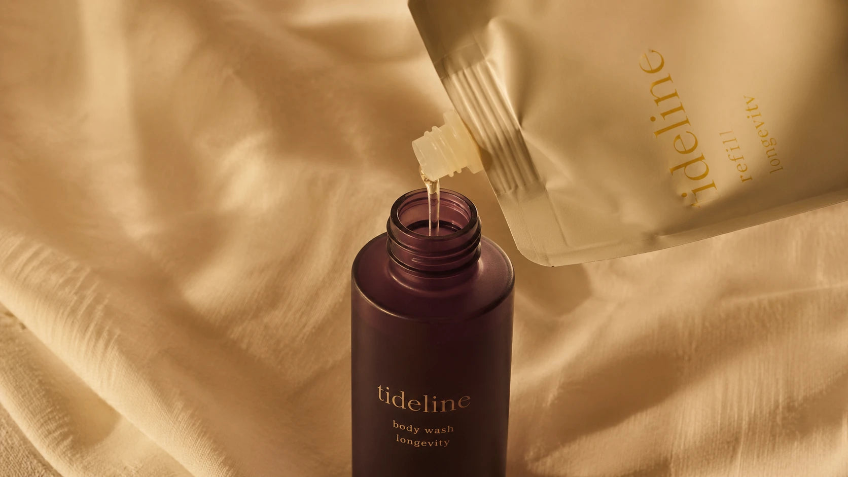 Macro detail of an aluminum refill pouch beside an empty Tideline glass primary bottle on bone-cream linen, soft tungsten light