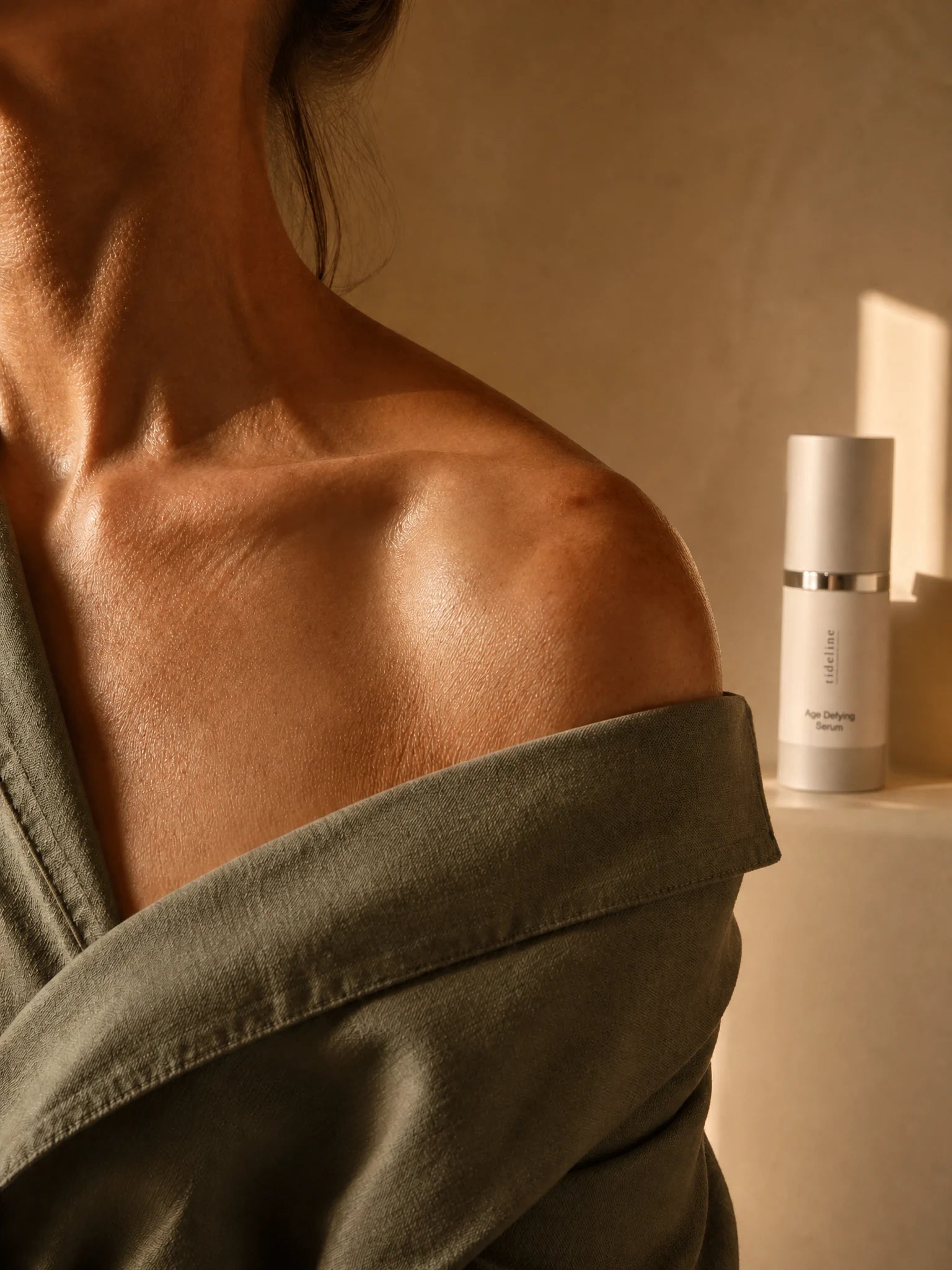 Neck, shoulder and décolleté of a woman in a soft linen robe in gentle window light, the Tideline Age-Defying Serum resting softly beside her